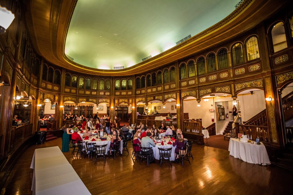 A large ornate wood-panelled hall has large arched windows, decorative details and a polished floor.