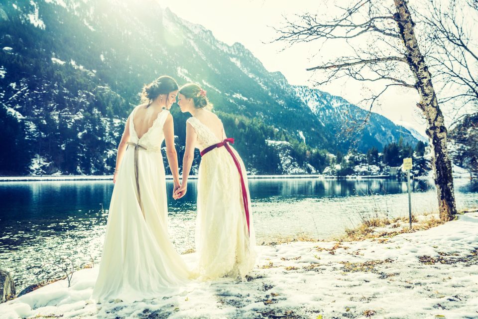 Dos mujeres con vestidos de novia caminan de la mano junto a un lago parcialmente helado.