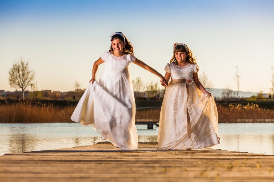 Dos niñas con vestidos blancos de comunión caminan de la mano sobre un embarcadero de madera junto al agua.