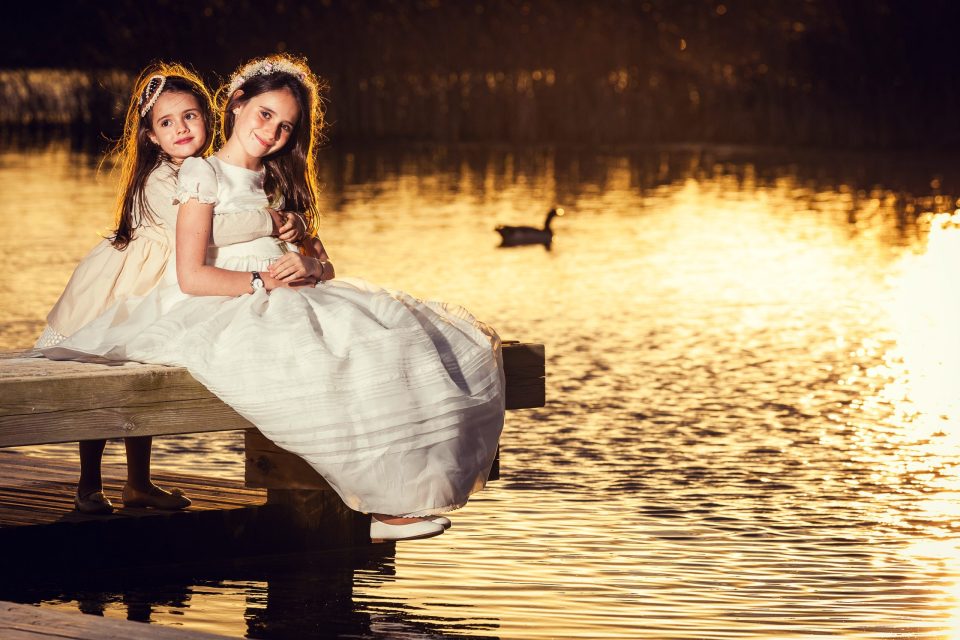 Dos niñas con vestidos blancos y adornos florales posan abrazadas sobre un embarcadero de madera junto a un lago.