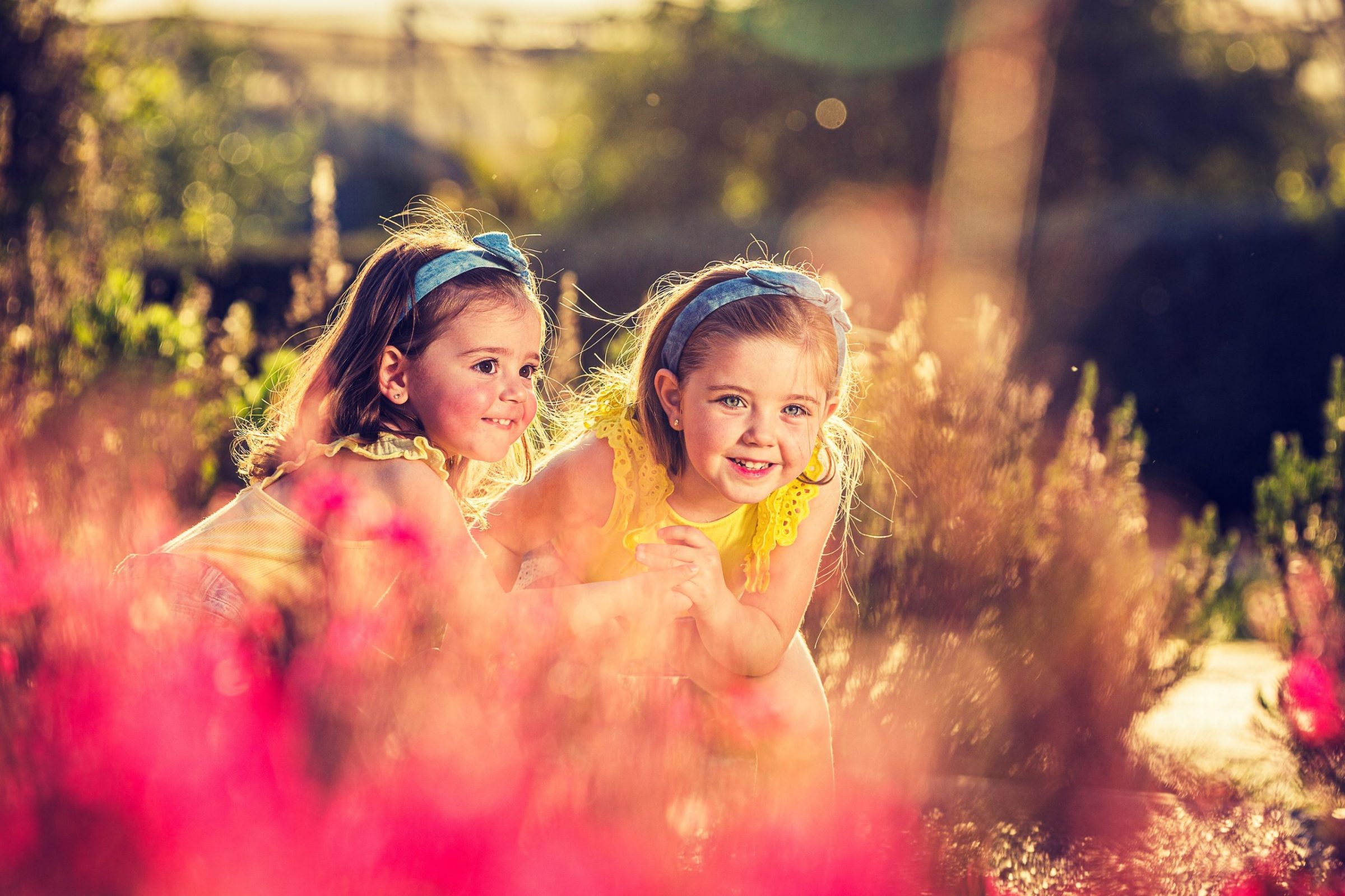 Dos niñas con vestidos y diademas se asoman entre flores en un parque arbolado.