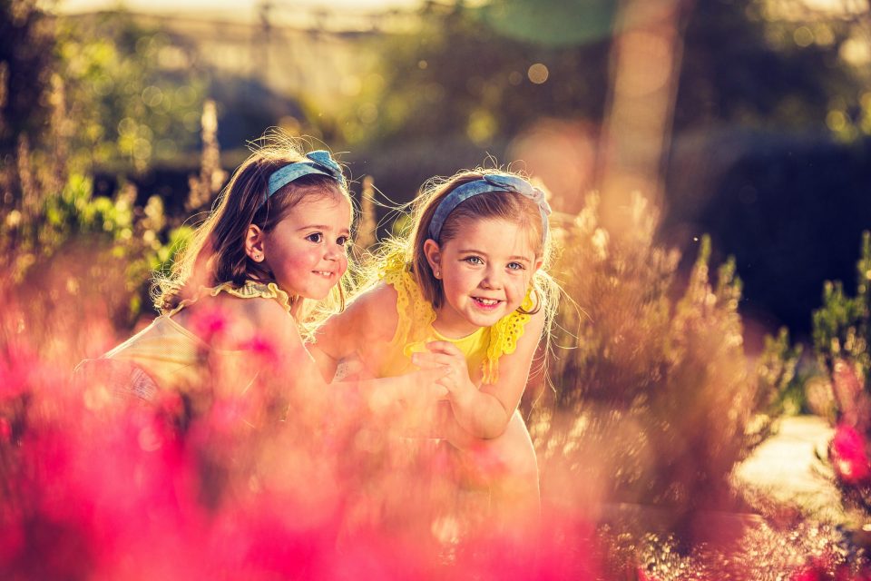 Dos niñas con vestidos y diademas se asoman entre flores en un parque arbolado.