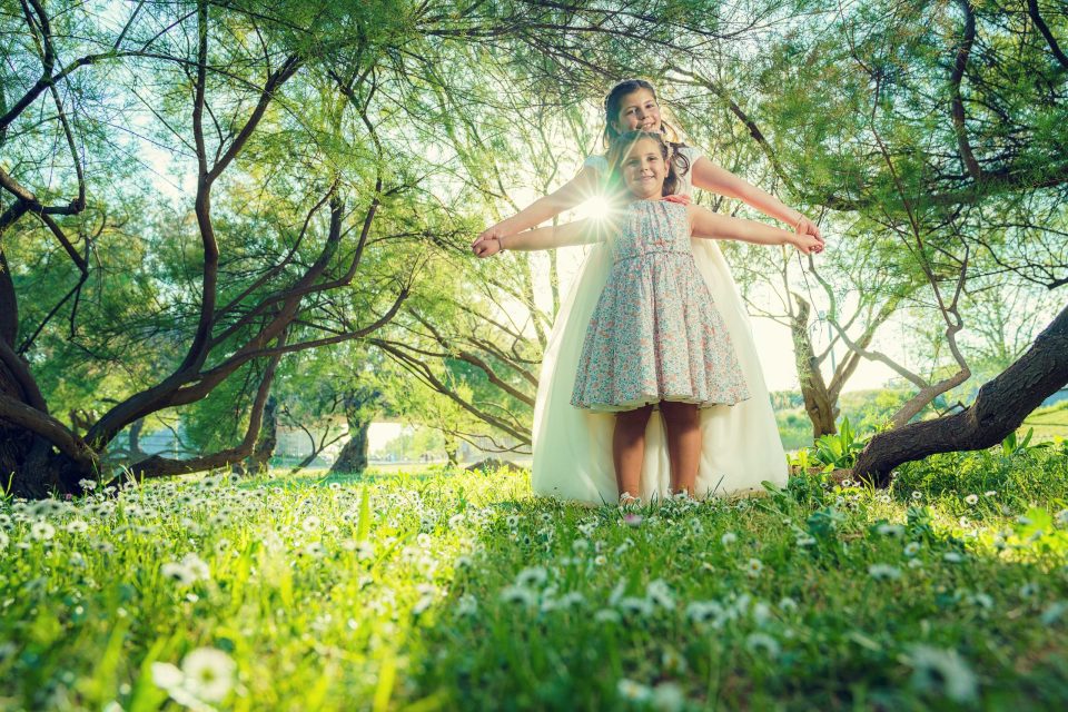 Dos niñas posan juntas en un parque cubierto de hierba y pequeñas flores blancas.