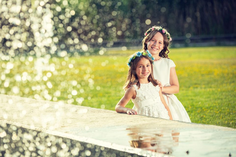 Dos niñas sonríen juntas en un parque, vestidas de blanco y con coronas de flores.