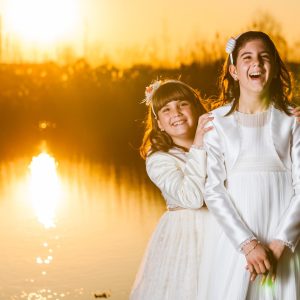 Two girls smile together beside a pond in a park, dressed in white for a First Communion session.