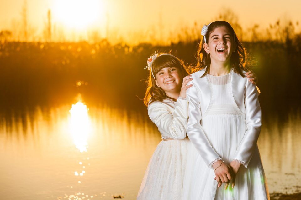 Dos niñas sonríen juntas junto a un estanque en un parque, vestidas de blanco para una sesión de comunión.