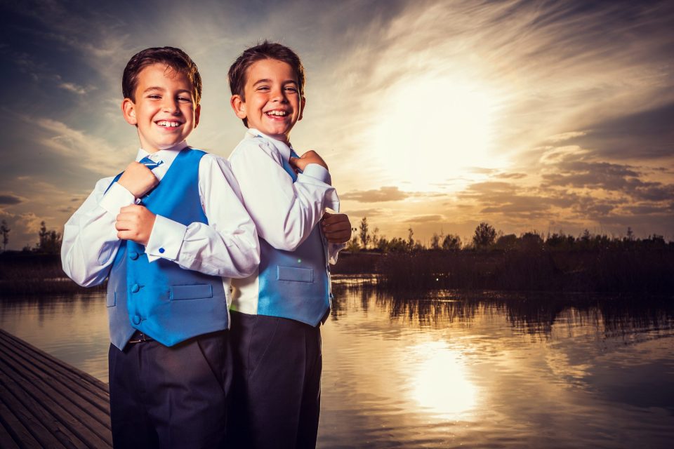 Dos niños sonrientes posan juntos con camisas blancas, corbatas y chalecos de tonos azul y gris.