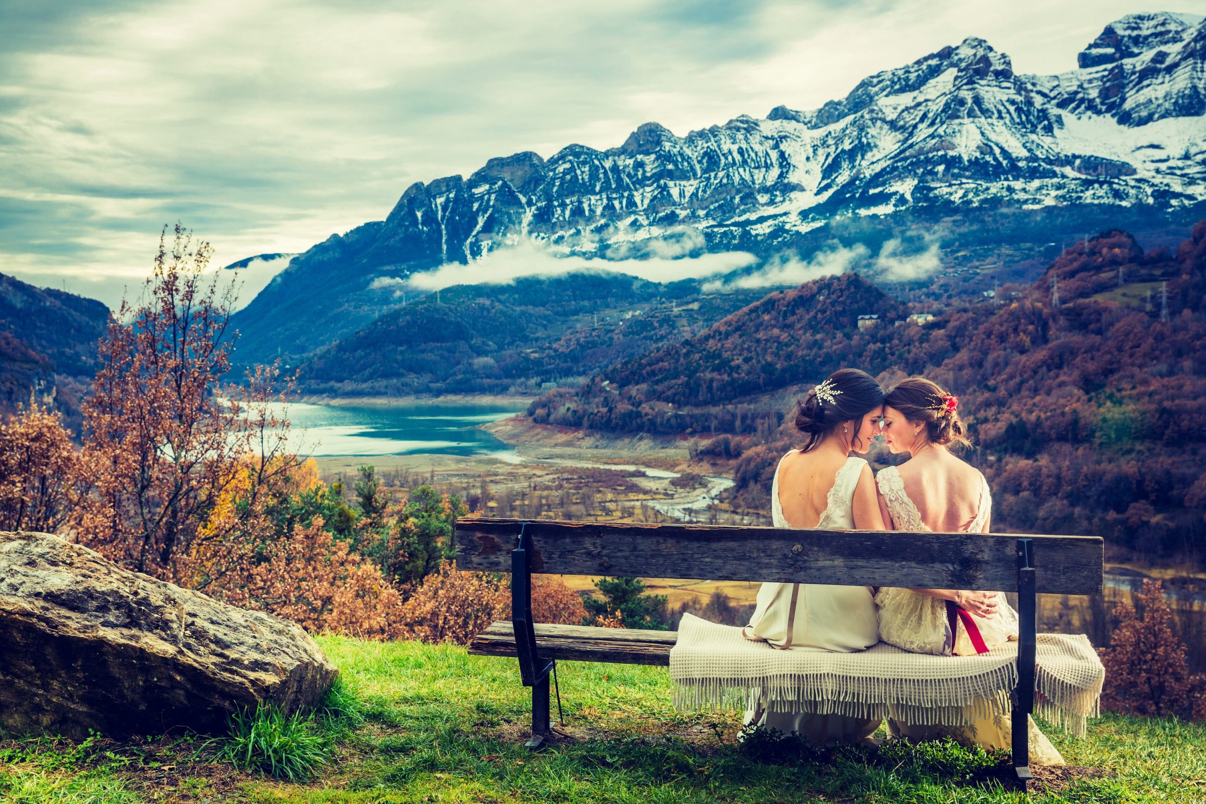 Two brides are sitting on a wooden bench, leaning in to kiss.