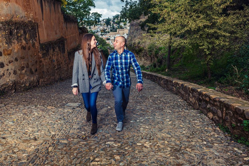 Two adults walk hand in hand and smile along a cobbled street lined by a stone wall and vegetation.