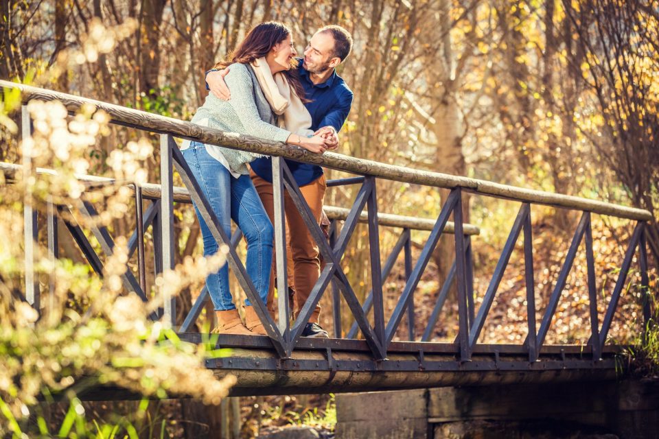 Two adults smile and embrace while standing on a wooden bridge with a dark railing.