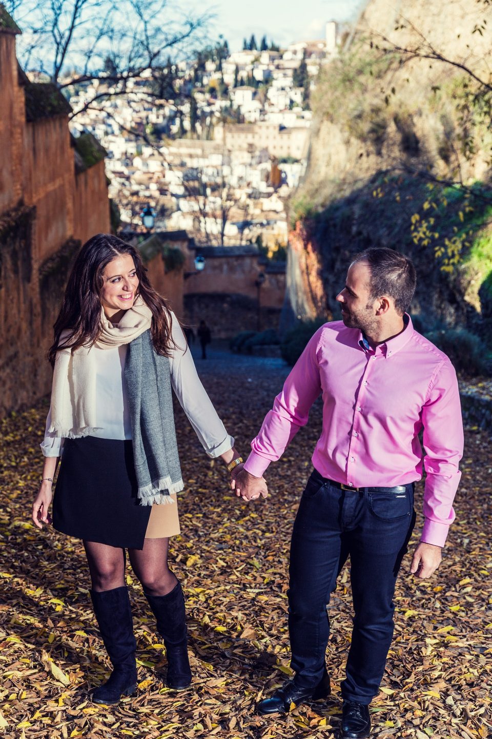 Two people walk hand in hand along a cobbled path covered with dry leaves.