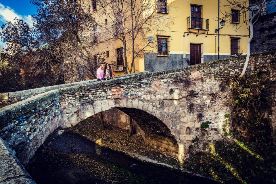 Two people walk across an old stone bridge over a narrow watercourse.