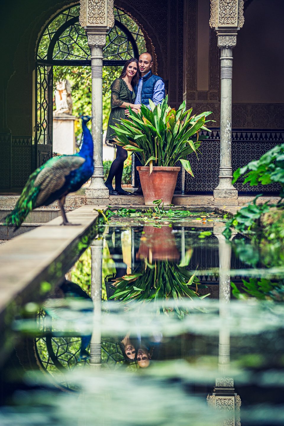 Two people pose together in a courtyard with ornate architecture, beside a large potted plant.