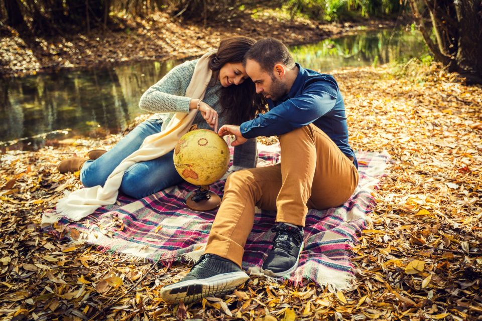 Two people are sitting together on a checked blanket in a wooded setting beside a pond.
