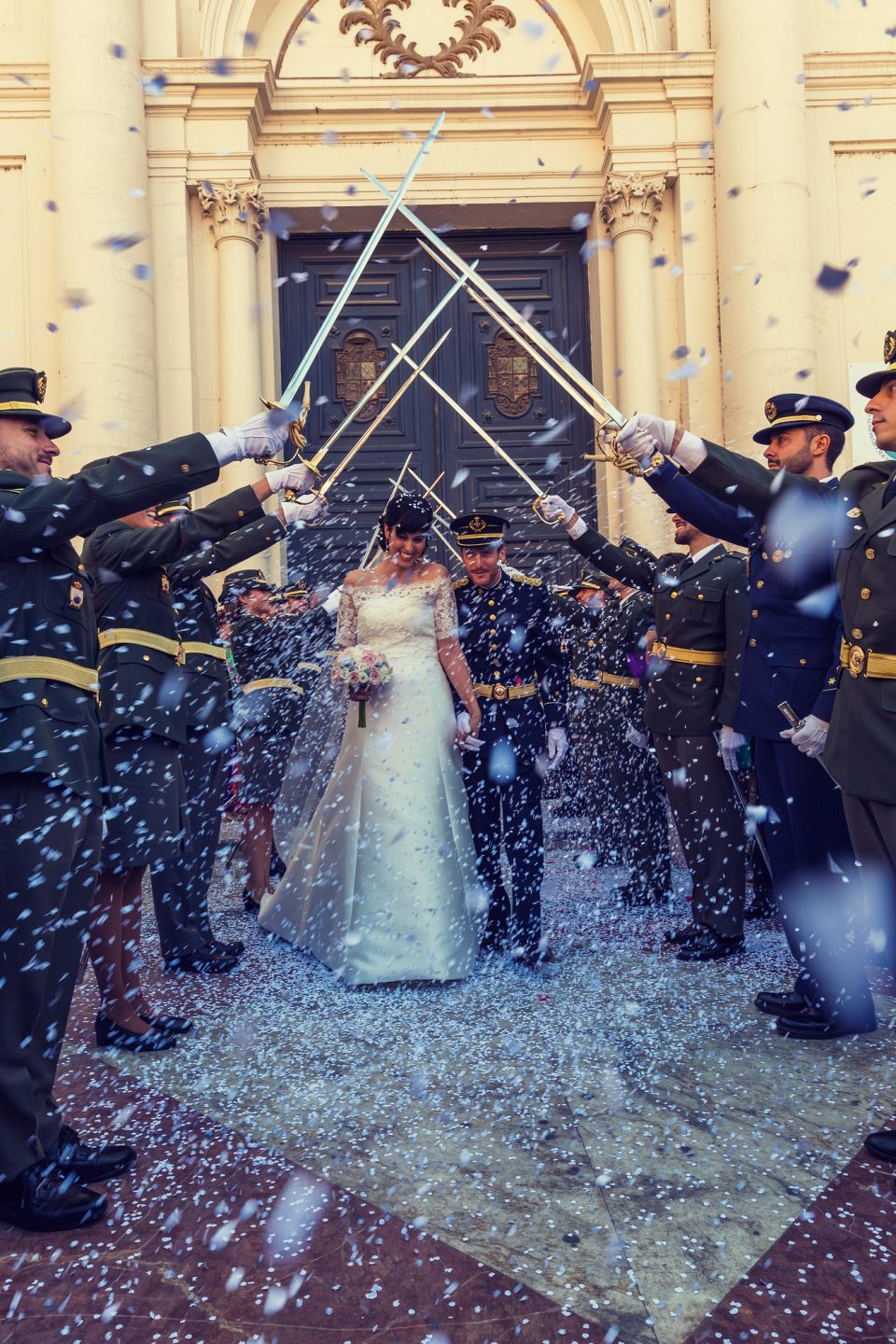 Escena de boda frente a una entrada monumental ornamentada: una novia con vestido blanco y ramo camina junto al novio.