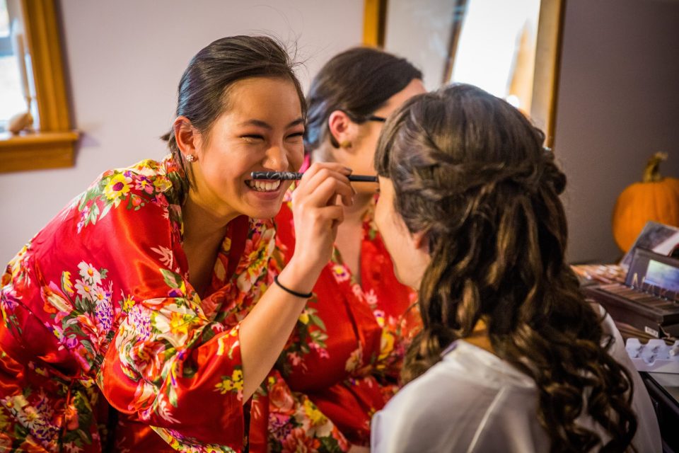 A woman smiles while applying eye make-up to another person during indoor wedding preparations.