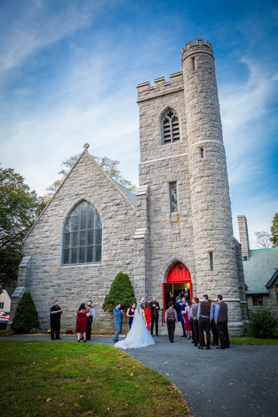 The exterior of a stone church features a circular battlemented tower, large windows and an open red door.