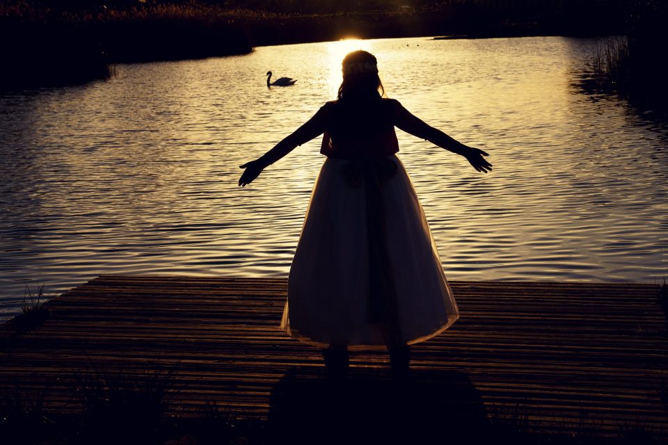 Fotografía a contraluz de una niña con un vestido claro, de pie sobre un embarcadero de madera en un parque.