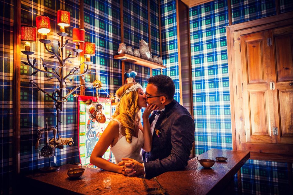Fotografía de boda en el interior de una finca: una pareja de novios se besa junto a una barra de madera.