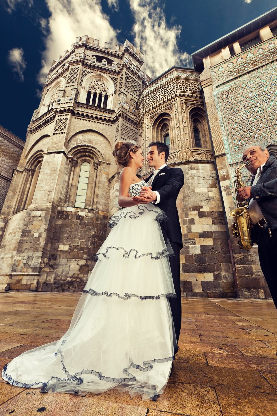 Fotografía de boda tomada desde un ángulo bajo frente a un monumento histórico de piedra ricamente ornamentado.