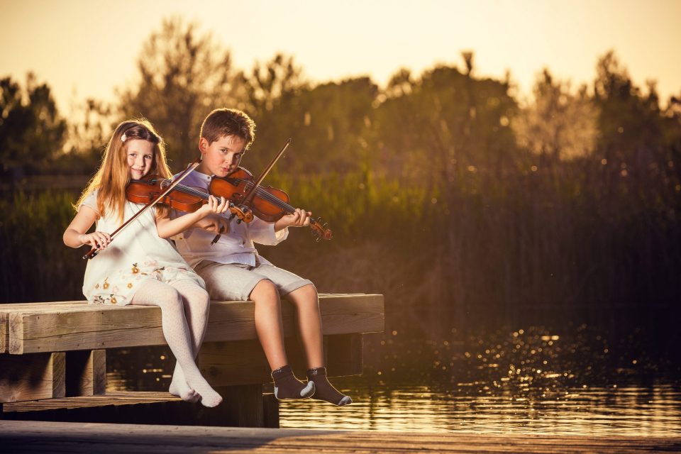 Fotografía de dos niños sentados en un muelle de madera sobre el agua, tocando el violín.