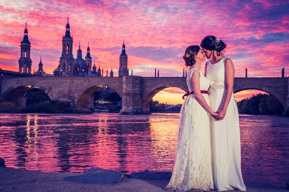 Fotografía de postboda al atardecer en una ciudad, con dos novias vestidas de blanco y abrazadas junto a un río.