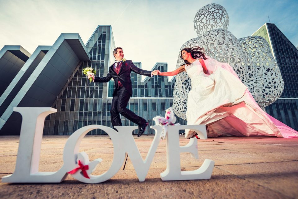 Fotografía de postboda en la que una pareja vestida para una boda corre mientras se toma de la mano frente a un edificio.