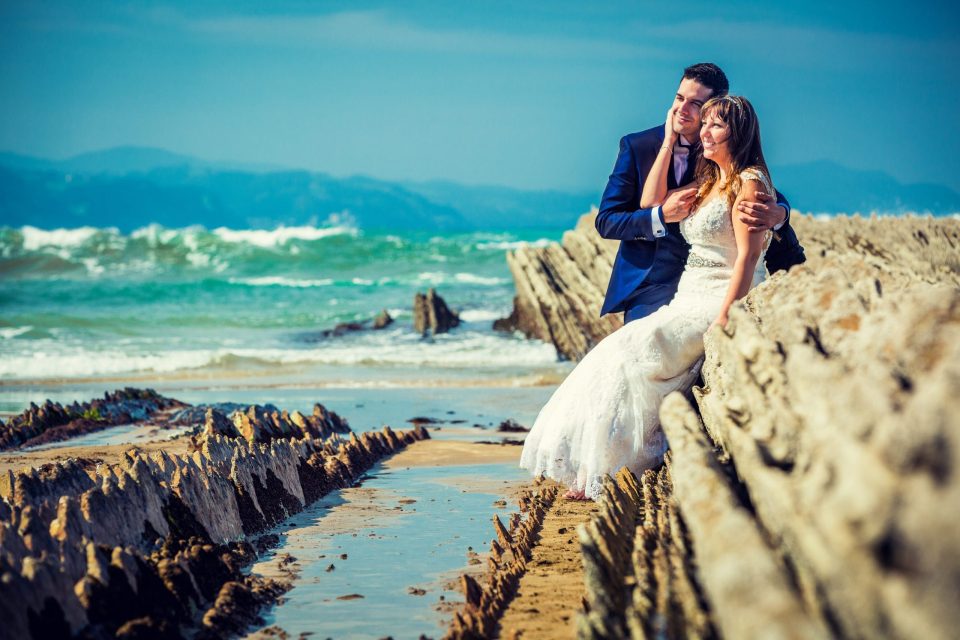 Fotografía de postboda en una playa rocosa: una pareja abraza y sonríe junto al mar.