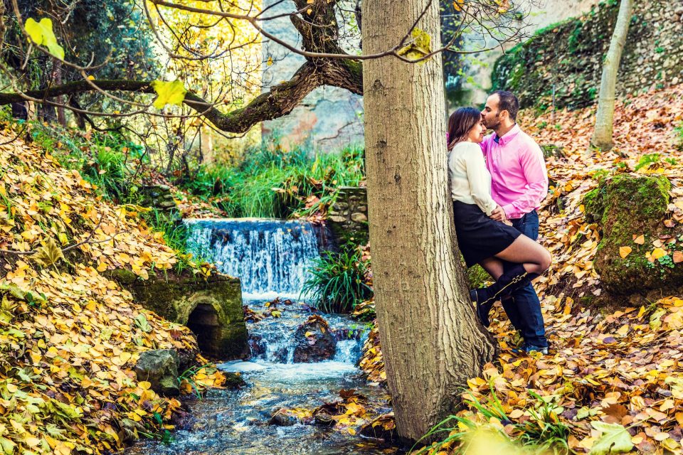 A couple embrace and kiss beside a tree in a wooded rural setting during autumn.