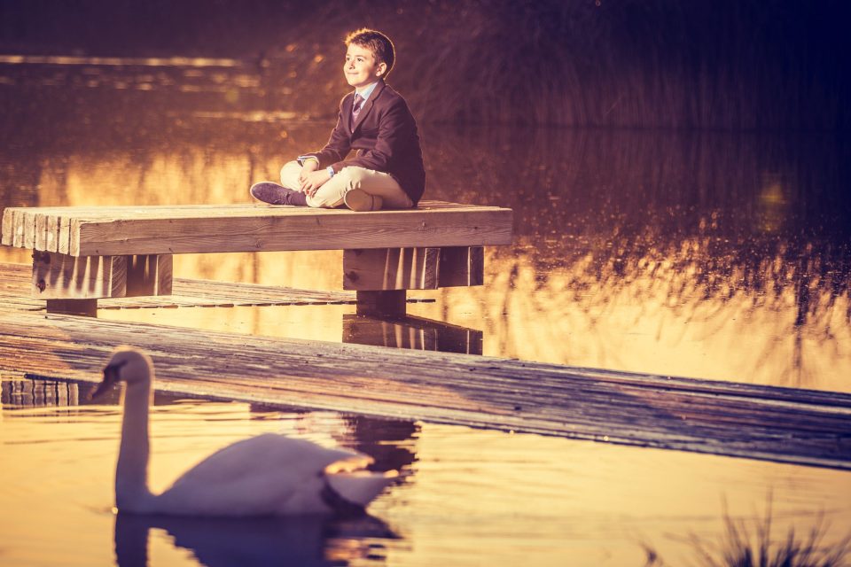 Fotografía de un niño vestido con traje oscuro, camisa y corbata, sentado sobre un embarcadero de madera junto a un lago.
