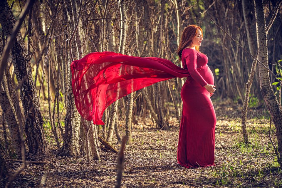Fotografía de una mujer embarazada con un vestido largo rojo en un parque arbolado.