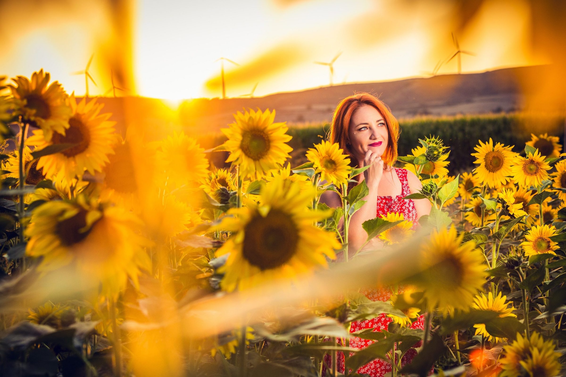 A red-haired woman in a patterned red dress is surrounded by flowering sunflowers.