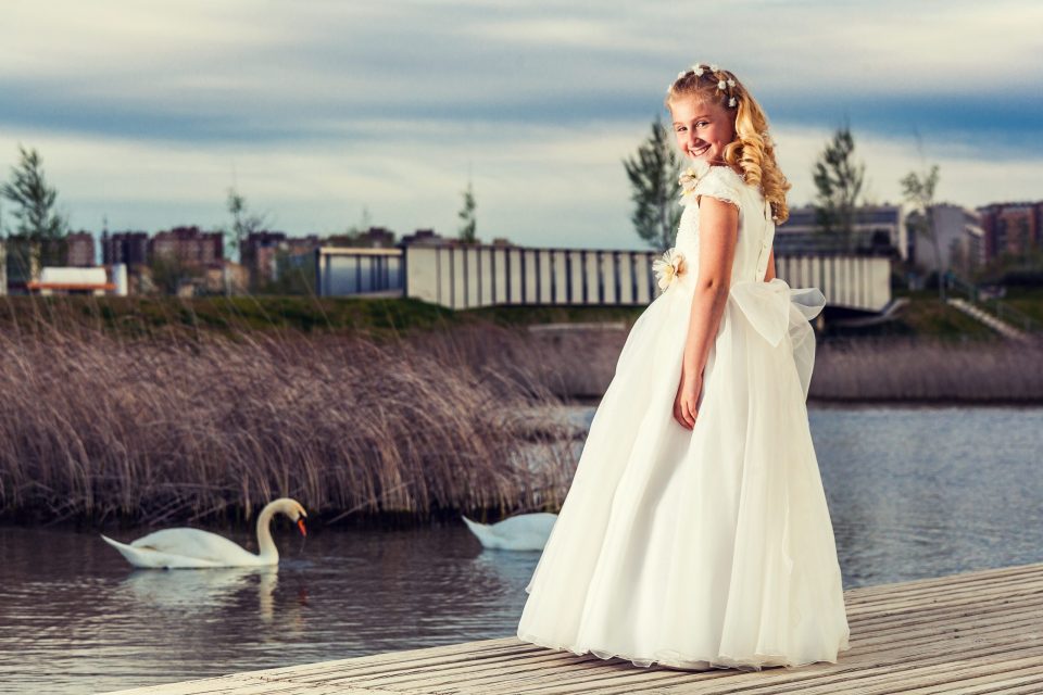 Fotografía de una niña con vestido blanco de comunión, decoración floral en el cabello y una gran lazada.
