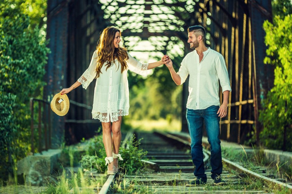 Fotografía de una pareja caminando y sonriendo sobre unas vías de tren rodeadas de vegetación.