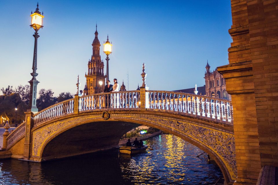 A bride and groom stand on an ornate bridge lit at dusk.