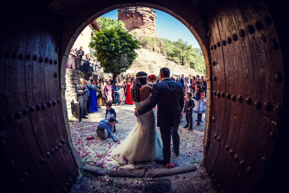 Fotografía de una pareja de novios vista de espaldas al salir de una iglesia, enmarcada por una gran puerta de madera.