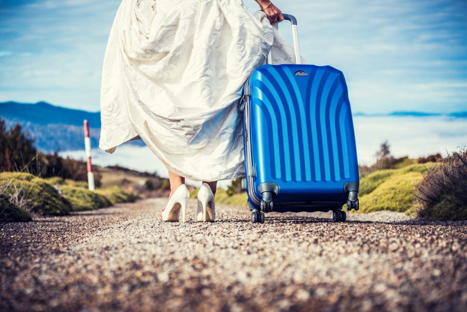 Fotografía de una persona con vestido blanco y zapatos de tacón que arrastra una maleta azul por un camino de grava.