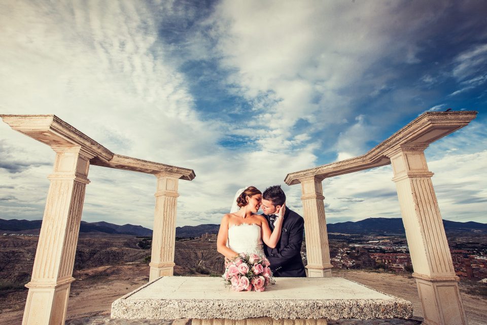 Fotografía exterior de una pareja de novios entre columnas de piedra de estilo clásico.