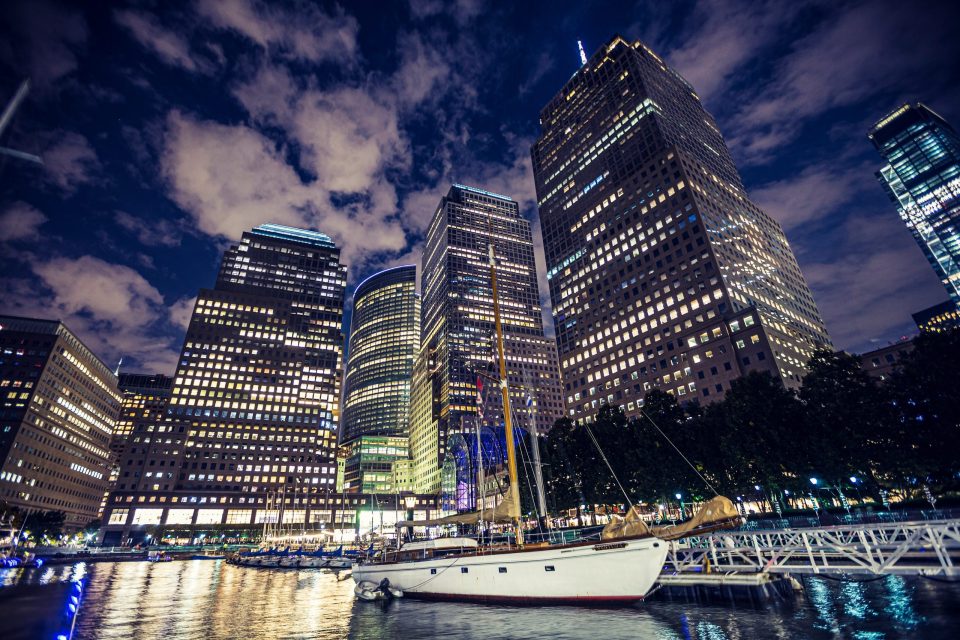 A white sailing boat is moored in the foreground of an urban harbour at night, with illuminated high-rise buildings in the background.
