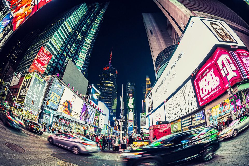A night-time photograph shows a broad urban intersection surrounded by tall buildings and large advertising screens.