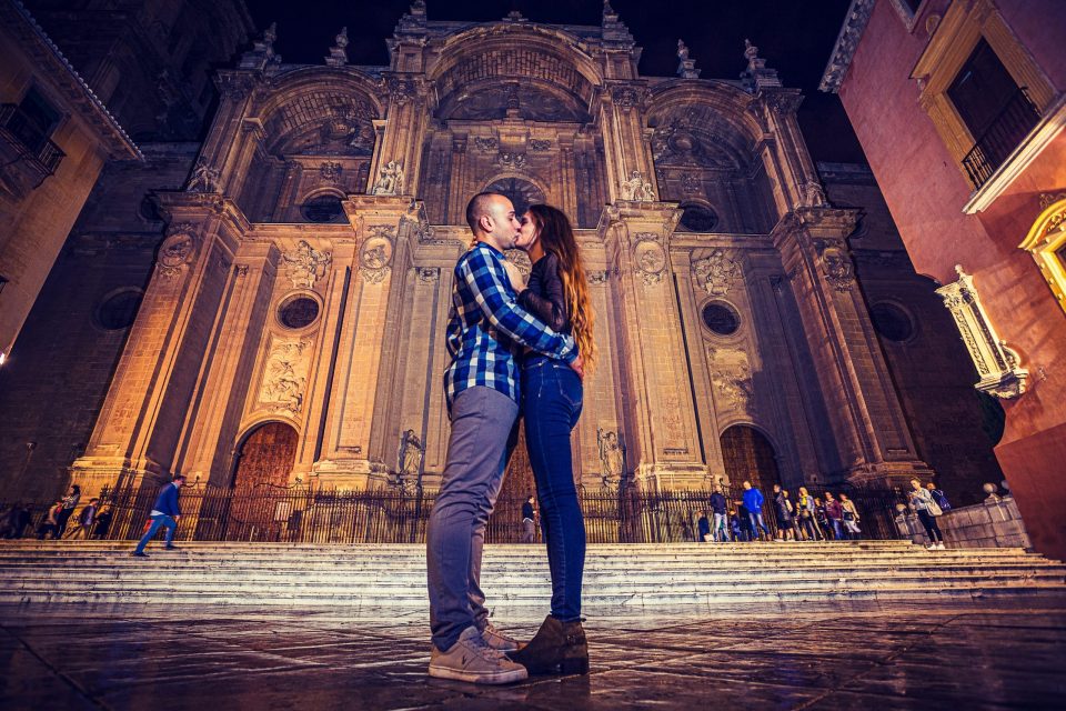 Fotografía nocturna de una pareja abrazada y besándose frente a la fachada iluminada de un monumento histórico.