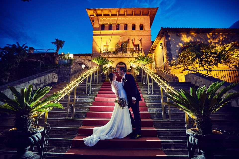 A bride and groom kiss on a red-carpeted staircase at night.