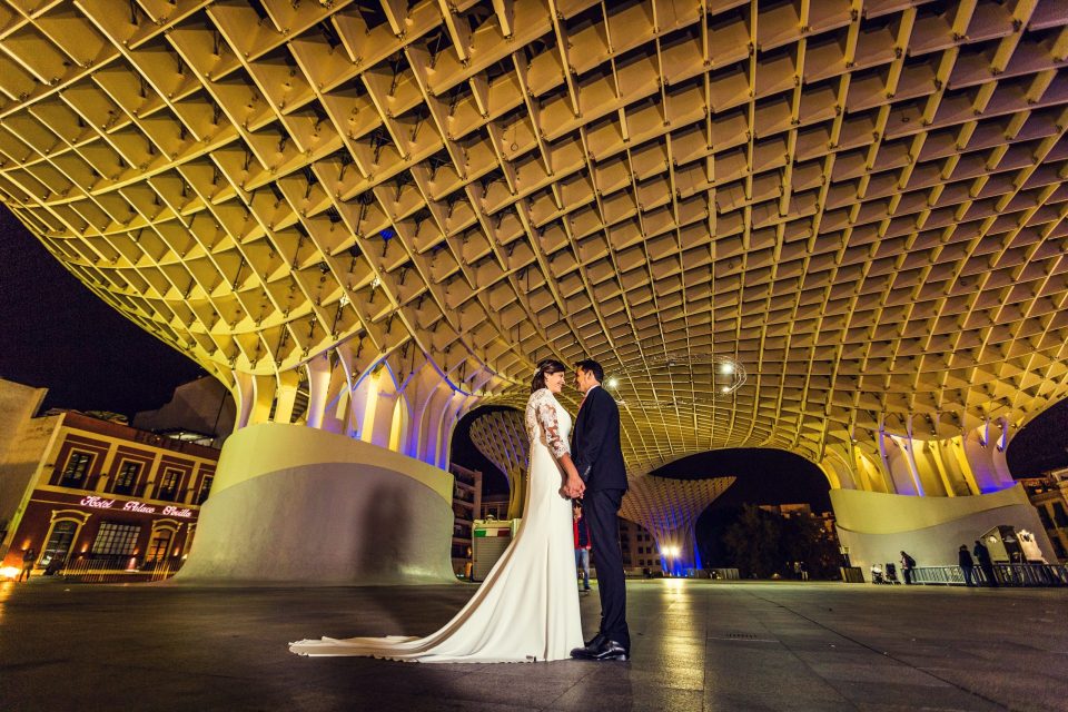 A newlywed couple stands beneath a large architectural structure with a geometric canopy at night.
