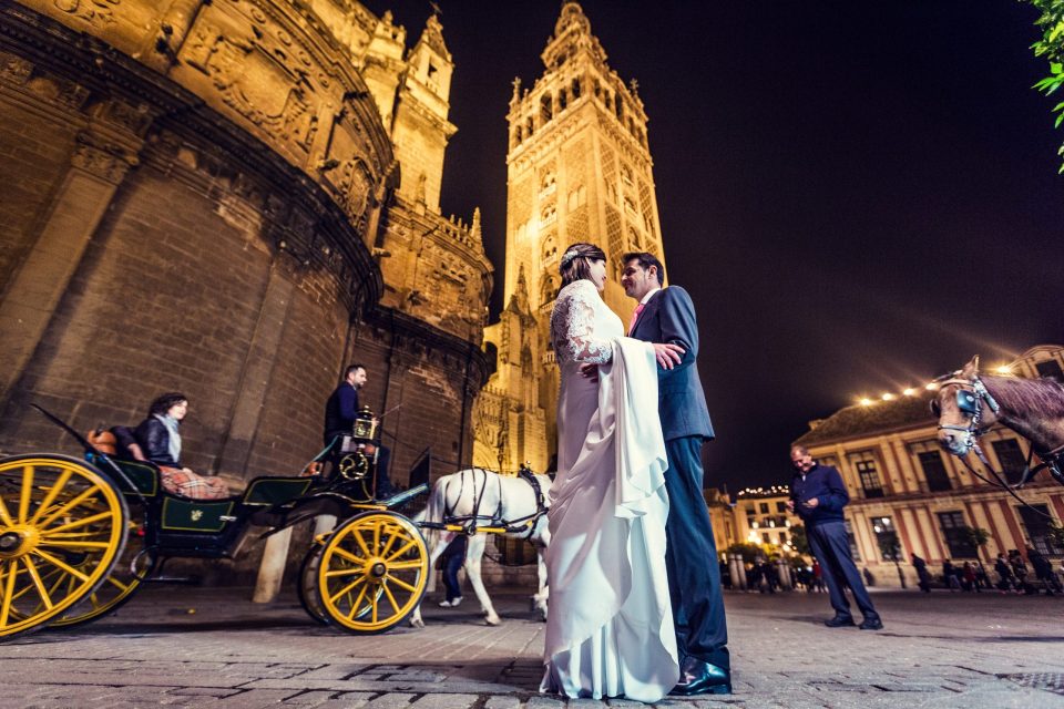 A wedding-dressed couple stands in front of an illuminated historic building at night.