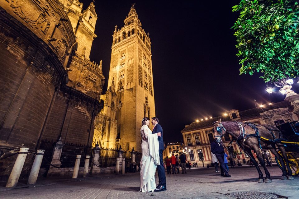 A couple dressed as bride and groom stand at night in front of an ornate church and an illuminated tower.