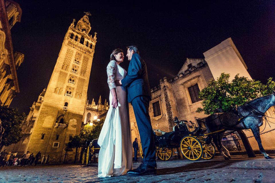 A couple dressed for a wedding stand in front of an illuminated historic tower at night.