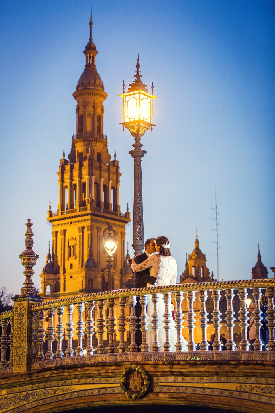 A couple kisses on an ornate bridge in a vertical sunset photograph.