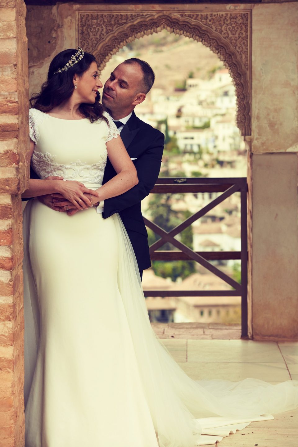 Fotografía vertical de una pareja de recién casados en actitud afectuosa, junto a un arco de piedra ornamentado.
