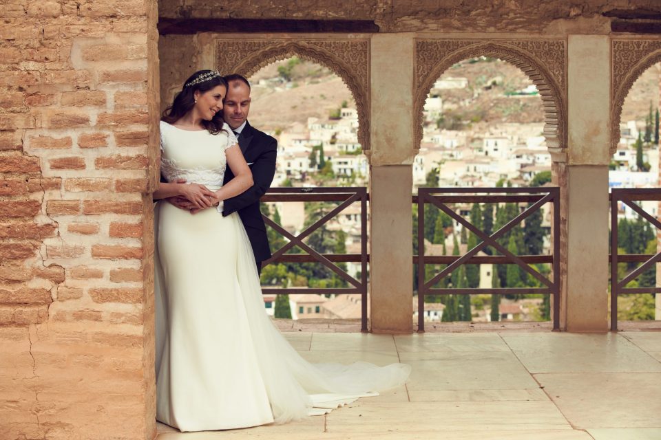 Fotos de Boda en la Alhambra de Granada. Una pareja de novios posa abrazada en una galería con arcos ornamentados.