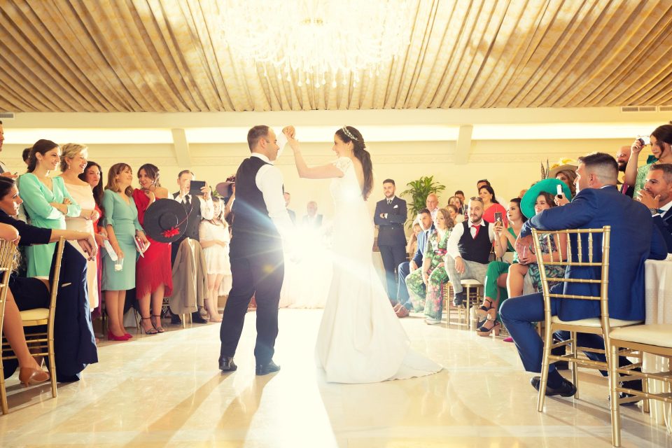Los novios bailan en el centro de un salón de una finca, rodeados de invitados que observan, sonríen y toman fotografías.